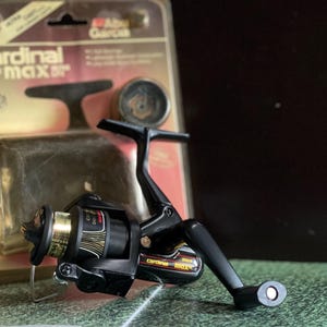 May include: Black and gold fishing reel with the text "Cardinal Max" on the side. The reel is in its original packaging, which is a clear plastic case with a cardboard backing. The packaging has the brand name "Abu Garcia" printed on it.
