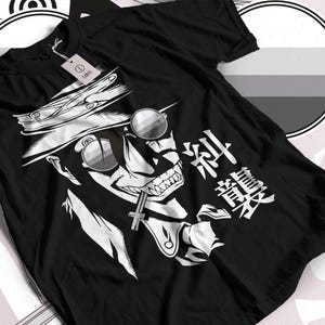 Puede incluir: Una camiseta negra de manga corta con un estampado gráfico blanco de un personaje con sombrero de ala ancha y gafas de sol redondas, con un objeto en forma de cruz en la boca y caracteres japoneses a la derecha de la cara.