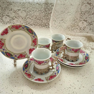 Peut inclure: Un ensemble de tasses et soucoupes vintage. Les tasses et soucoupes présentent un motif floral avec des accents rouges, roses et bleus. Les tasses ont des poignées en métal ornées et sont posées sur une surface blanche.