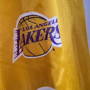 Pu&ograve; includere: Primo piano di una maglia da basket gialla con il logo dei Los Angeles Lakers. Il logo presenta un pallone da basket con le parole "Los Angeles" sopra e "Lakers" in viola sotto. La maglia ha un tessuto strutturato.