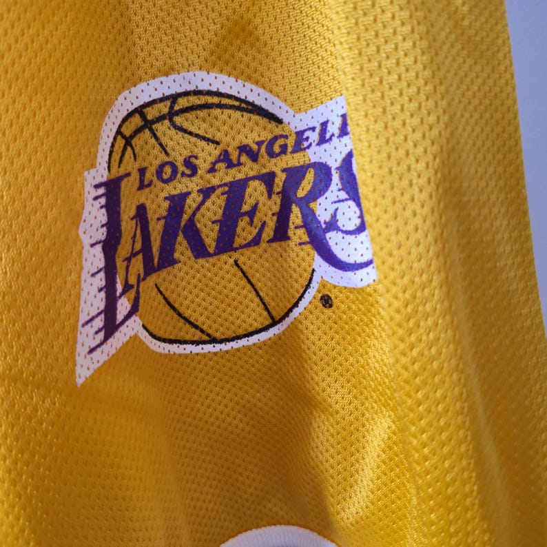 Pu&ograve; includere: Primo piano di una maglia da basket gialla con il logo dei Los Angeles Lakers. Il logo presenta un pallone da basket con le parole "Los Angeles" sopra e "Lakers" in viola sotto. La maglia ha un tessuto strutturato.