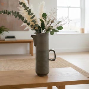May include: A grey ceramic vase with a handle and spout, holding dried flowers and greenery. The vase has a textured lower section and is placed on a wooden table. The arrangement includes pampas grass, eucalyptus, and other decorative elements.