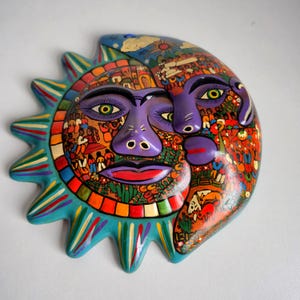 Könnte beinhalten: Eine farbenfrohe, handgefertigte Sonnen- und Mondmaske. Die Maske hat ein lila Gesicht mit grünen Augen und roten Lippen. Die Sonnenstrahlen sind türkis mit bunten Streifen. Die Maske ist wahrscheinlich aus Keramik oder einem ähnlichen Material.