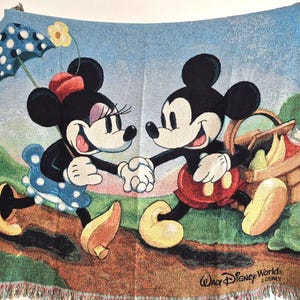May include: A woven throw blanket featuring Mickey and Minnie Mouse holding hands, with a picnic basket and umbrella. The blanket has a light blue background with a fringed edge and the text "Walt Disney World" at the bottom.
