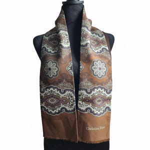 May include: A brown silk scarf with a repeating floral and paisley pattern in shades of white, brown, and purple. The scarf is draped over a black mannequin and features the text "Christian Dior" in white.