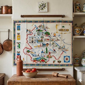 Könnte beinhalten: Eine Vintage-Karte von Abruzzen, Italien, auf einem weißen Tuch mit farbenfrohem Rand. Die Karte ist detailliert mit Illustrationen von Sehenswürdigkeiten und Figuren. Eine Keramikflasche und eine Schüssel mit Tomaten stehen auf einer Holzoberfläche.