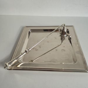 Peut inclure: Un plateau carré en métal argenté poli avec un bord surélevé et une surface lisse et réfléchissante. Une longue poignée métallique fine, dotée d'un petit embout en forme de cloche à une extrémité, repose en diagonale sur le plateau.