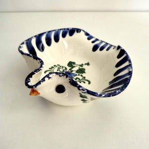 May include: A cream-coloured ceramic bowl shaped like a bird with a scalloped rim painted in dark blue. The interior features a green and blue floral vine pattern. The bird has a small orange beak and a dark blue eye, with blue sponged edges.