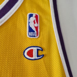 Pu&ograve; includere: Primo piano di una maglia da basket gialla con finiture viola e bianche. Presenta il logo NBA e una toppa con il logo Champion. La maglia ha un tessuto strutturato e traspirante.
