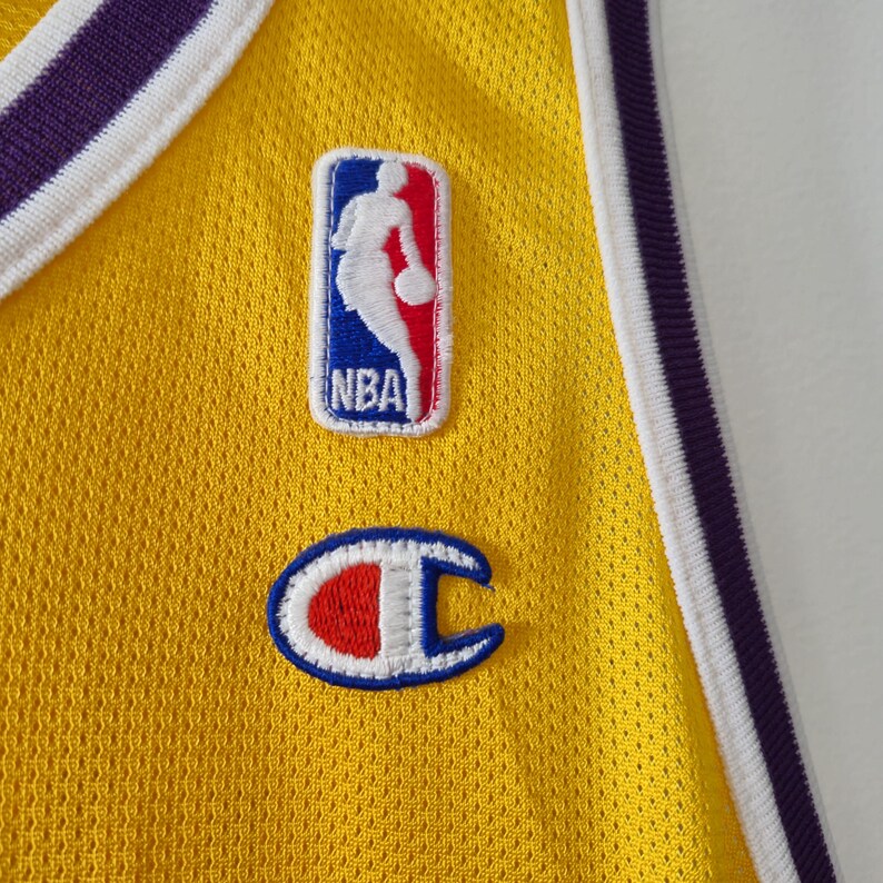 Pu&ograve; includere: Primo piano di una maglia da basket gialla con finiture viola e bianche. Presenta il logo NBA e una toppa con il logo Champion. La maglia ha un tessuto strutturato e traspirante.