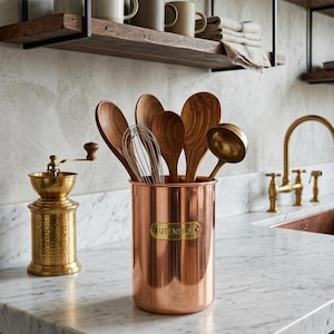 May include: A copper utensil holder with a brass label that reads "UTENSILS" contains wooden spoons, a whisk, and a ladle. A brass coffee grinder sits to the left. The kitchen setting includes a marble countertop and wooden shelves.