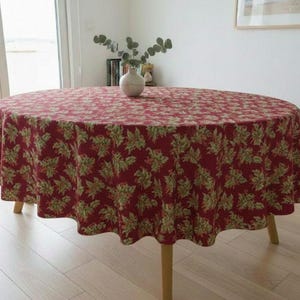 May include: A round tablecloth featuring a deep red background with a repeating pattern of green holly leaves and small red berries. The fabric drapes over a circular table with wooden legs, creating a consistent textile covering across the surface.