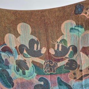 May include: A woven textile featuring Mickey and Minnie Mouse in a colorful design. The artwork includes the iconic characters in a light color, set against a brown background with floral and abstract elements in shades of blue, green, and peach.