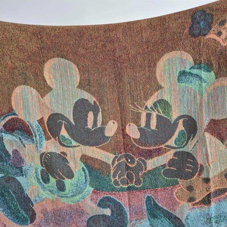May include: A woven textile featuring Mickey and Minnie Mouse in a colorful design. The artwork includes the iconic characters in a light color, set against a brown background with floral and abstract elements in shades of blue, green, and peach.