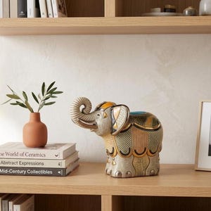 May include: A detailed elephant ornament in grey, gold, and green, with a raised trunk. It sits on a wooden shelf alongside a terracotta vase with greenery and a stack of books. The ornament is a decorative item.