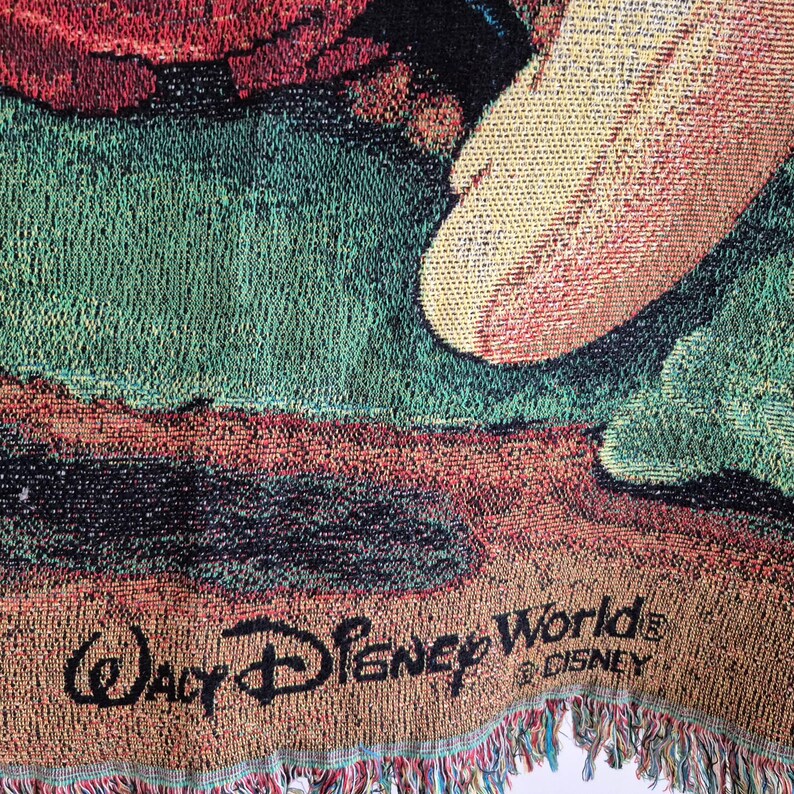 May include: Woven tapestry featuring a colorful design with the words "Walt Disney World" in black. The tapestry has a fringed edge and a mix of green, red, and tan hues.