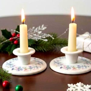May include: Two white ceramic candle holders with a floral pattern around the base, each holding a lit cream-colored taper candle. The candle holders feature a wide, flat circular base and a central cup to secure the candle in an upright position.