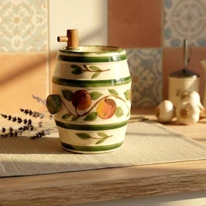 May include: A ceramic barrel-shaped container with a wooden spigot. The off-white container is decorated with green stripes and a design of peaches and leaves. The container is sitting on a beige placemat.