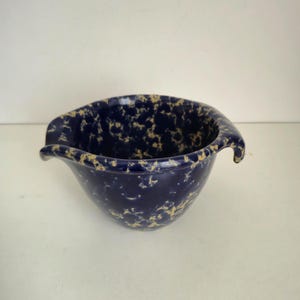 May include: A navy blue ceramic mixing bowl with a speckled tan pattern. The bowl has a pouring spout and a handle on opposite sides. This vintage kitchenware item is ideal for baking and cooking.