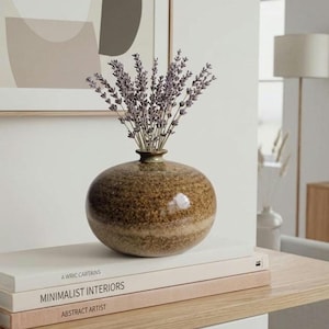 May include: A round, speckled brown ceramic vase with a short neck, containing a bouquet of dried purple lavender stems. The vase rests on a stack of three books with visible text including "Minimalist Interiors" and "Abstract Artist."