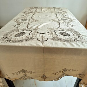 May include: Beige rectangular tablecloth with detailed embroidered floral and geometric patterns. The edges have a scalloped design with intricate embroidery. The tablecloth is draped over a dark surface.