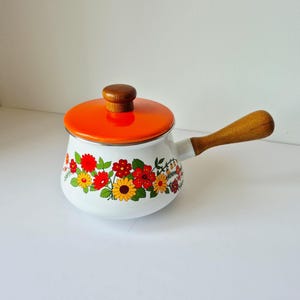 May include: White enamel saucepan with an orange lid and wooden handle. The pot is decorated with a floral pattern of red, yellow, and orange flowers with green leaves. The lid has a wooden knob.