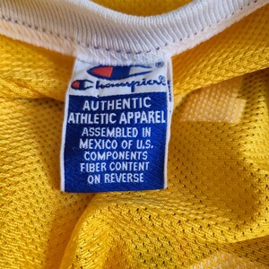 Pu&ograve; includere: Primo piano di una maglia sportiva in rete gialla con colletto bianco. &Egrave; visibile un'etichetta blu con il logo Champion e la scritta "AUTHENTIC ATHLETIC APPAREL". L'etichetta indica anche "ASSEMBLED IN MEXICO OF U.S. COMPONENTS".