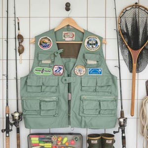 Peut inclure: Un gilet de pêche vert clair avec plusieurs poches et écussons, suspendu à un cintre en bois. Des cannes à pêche, une épuisette et une boîte à pêche avec des leurres sont également visibles, suggérant un thème de pêche.