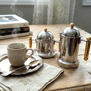 May include: A coffee set featuring a silver tray, cup, and spoon, with two stainless steel coffee pots with gold handles. A stack of books is in the background. The scene is set on a wooden table with a linen napkin.