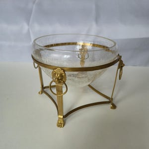 May include: A clear crackle glass bowl set within a gold-toned metal stand. The stand features four fluted legs ending in paw-shaped feet, with each leg topped by a lion head ornament holding a circular ring in its mouth, supporting the bowl.