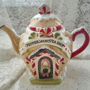 May include: A novelty ceramic teapot designed as a gingerbread house. It has a tan base with white icing, red and white candy cane decorations, and the words "Peppermint Tea Shop". The handle is red.