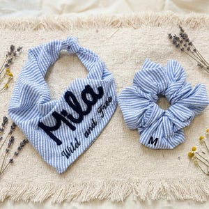 May include: A blue and white pinstriped fabric bandana featuring the embroidered black text "Mila" and "Wild and Free," paired with a matching blue and white pinstriped fabric hair scrunchie that has the letter "M" printed on it.