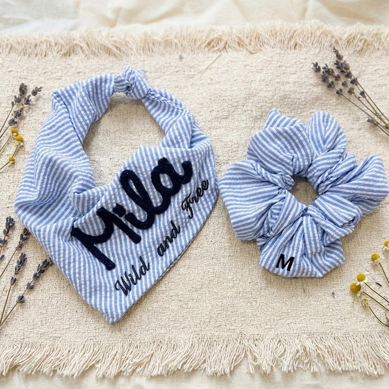 May include: A blue and white pinstriped fabric bandana featuring the embroidered black text "Mila" and "Wild and Free," paired with a matching blue and white pinstriped fabric hair scrunchie that has the letter "M" printed on it.