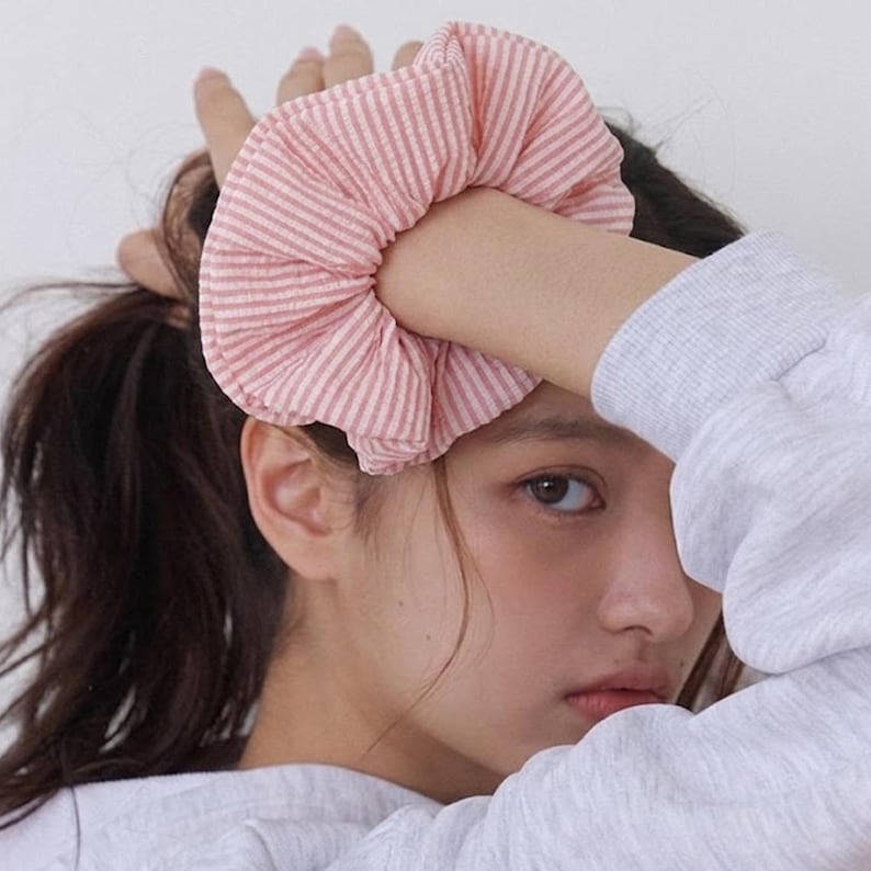 May include: A fabric hair scrunchie featuring a light pink and white pinstripe pattern. The accessory is gathered into a circular, ruffled shape with a soft, textured appearance, designed to hold hair in place with its elasticized interior construction.