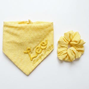 May include: A yellow and white seersucker fabric bandana featuring the embroidered text "Lee" in a cursive font, accompanied by the smaller text "Always Happy" below it, paired with a matching yellow and white striped fabric hair scrunchie.