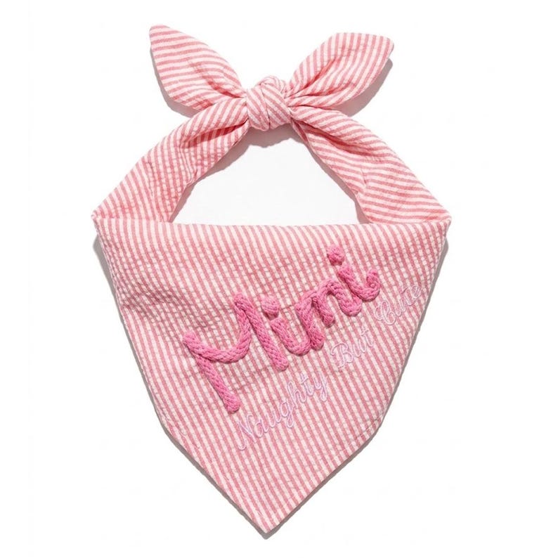 May include: A pink and white striped triangular pet bandana featuring a knotted top. The word Mimi is embroidered in large pink script, with the text Naughty But Cute embroidered in smaller pink script directly underneath the main name.