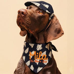 May include: A dark navy blue pet bandana and matching baseball cap featuring a repeating white ghost pattern. The bandana is embroidered with the name Mila in orange script, followed by the text Too Cute to Spook in smaller orange lettering.