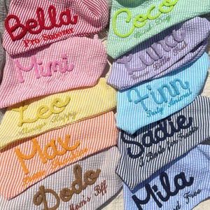 May include: A collection of triangular pet bandanas made from seersucker fabric in various colors, each featuring embroidered names and phrases such as Bella Pro Snoozer, Coco Good Dog, Mimi, Lulu Brave Little Soul, Finn Party Animal, and Sadie I Bark for Treats.