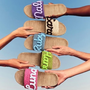 K&ouml;nnte beinhalten: F&uuml;nf Pantoletten mit geflochtenen beigen Sohlen und Riemen in Lila, Beige, Blau, Gr&uuml;n und Pink. Jeder Riemen zeigt einen Namen in Schreibschrift: Nala, Sara, Ruby, Hana und Luna, gearbeitet aus kontrastierendem wei&szlig;em, braunem oder helllila Garn.