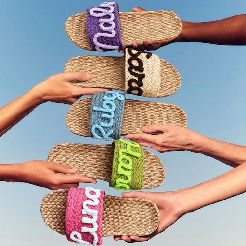 K&ouml;nnte beinhalten: F&uuml;nf Pantoletten mit geflochtenen beigen Sohlen und Riemen in Lila, Beige, Blau, Gr&uuml;n und Pink. Jeder Riemen zeigt einen Namen in Schreibschrift: Nala, Sara, Ruby, Hana und Luna, gearbeitet aus kontrastierendem wei&szlig;em, braunem oder helllila Garn.