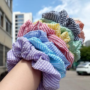 May include: A stack of multiple fabric hair scrunchies featuring a seersucker texture with thin white stripes. The scrunchies are arranged in a rainbow color gradient, including shades of purple, blue, light blue, red, yellow, green, and black.