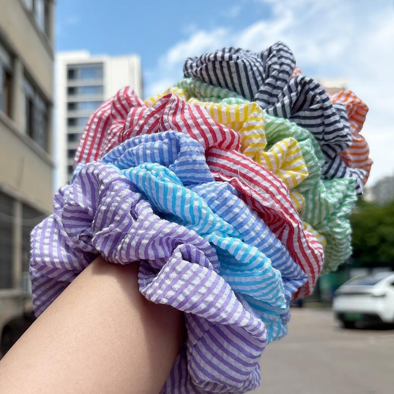 May include: A stack of multiple fabric hair scrunchies featuring a seersucker texture with thin white stripes. The scrunchies are arranged in a rainbow color gradient, including shades of purple, blue, light blue, red, yellow, green, and black.