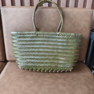 May include: A green woven tote bag with braided handles. The bag has a rectangular shape and a textured, chevron-like pattern. The bag is made of a natural material and is sitting on a brown surface.
