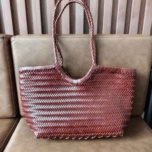 Può includere: Una borsa tote marrone intrecciata con manici intrecciati. La borsa ha una forma rettangolare con un'apertura curva e un motivo a trama fitta. I manici sono anch'essi marroni e intrecciati, offrendo un design robusto ed elegante.