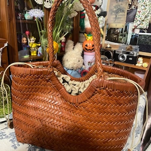 K&ouml;nnte beinhalten: Eine braune, gewebte Leder-Tragetasche mit geflochtenem Griff. Die Tasche hat ein einzigartiges offenes Design und ist mit einem hellfarbigen Stoff gef&uuml;ttert. Ein stilvolles Accessoire zum Tragen von Gegenst&auml;nden.