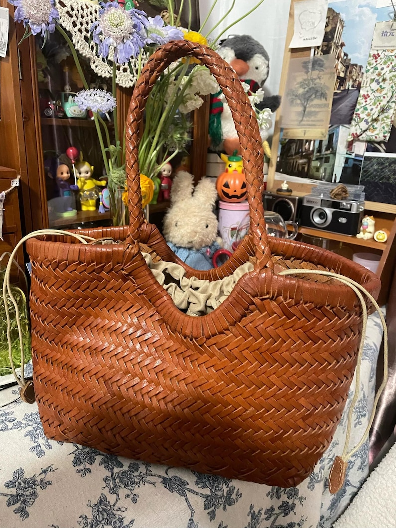 K&ouml;nnte beinhalten: Eine braune, gewebte Leder-Tragetasche mit geflochtenem Griff. Die Tasche hat ein einzigartiges offenes Design und ist mit einem hellfarbigen Stoff gef&uuml;ttert. Ein stilvolles Accessoire zum Tragen von Gegenst&auml;nden.