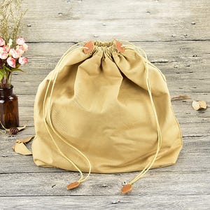 K&ouml;nnte beinhalten: Ein beigefarbener Kordelzugbeutel mit geraffter Oberseite und lederartigen Akzenten. Die Tasche besteht aus einem weichen, strapazierf&auml;higen Stoff und verf&uuml;gt &uuml;ber einen Kordelzugverschluss. Die Tasche liegt auf einer Holzoberfl&auml;che.