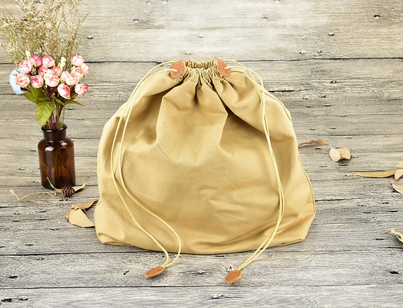 K&ouml;nnte beinhalten: Ein beigefarbener Kordelzugbeutel mit geraffter Oberseite und lederartigen Akzenten. Die Tasche besteht aus einem weichen, strapazierf&auml;higen Stoff und verf&uuml;gt &uuml;ber einen Kordelzugverschluss. Die Tasche liegt auf einer Holzoberfl&auml;che.