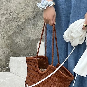K&ouml;nnte beinhalten: Eine braune, gewebte Tragetasche mit geflochtenen Henkeln wird von einer Person getragen, die ein blaues Jeanskleid tr&auml;gt. Ein wei&szlig;es Scrunchie mit floralem Muster und ein goldener Ring sind sichtbar. Die Tasche hat eine rechteckige Form und ein gewebtes Design.