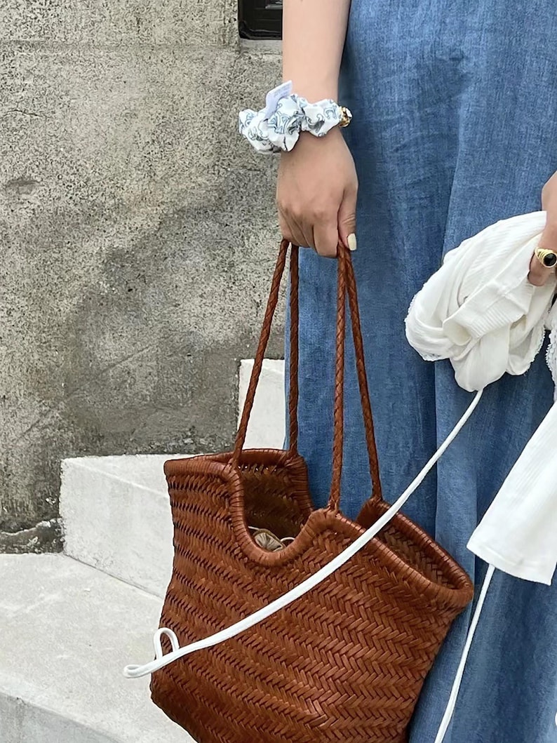 K&ouml;nnte beinhalten: Eine braune, gewebte Tragetasche mit geflochtenen Henkeln wird von einer Person getragen, die ein blaues Jeanskleid tr&auml;gt. Ein wei&szlig;es Scrunchie mit floralem Muster und ein goldener Ring sind sichtbar. Die Tasche hat eine rechteckige Form und ein gewebtes Design.